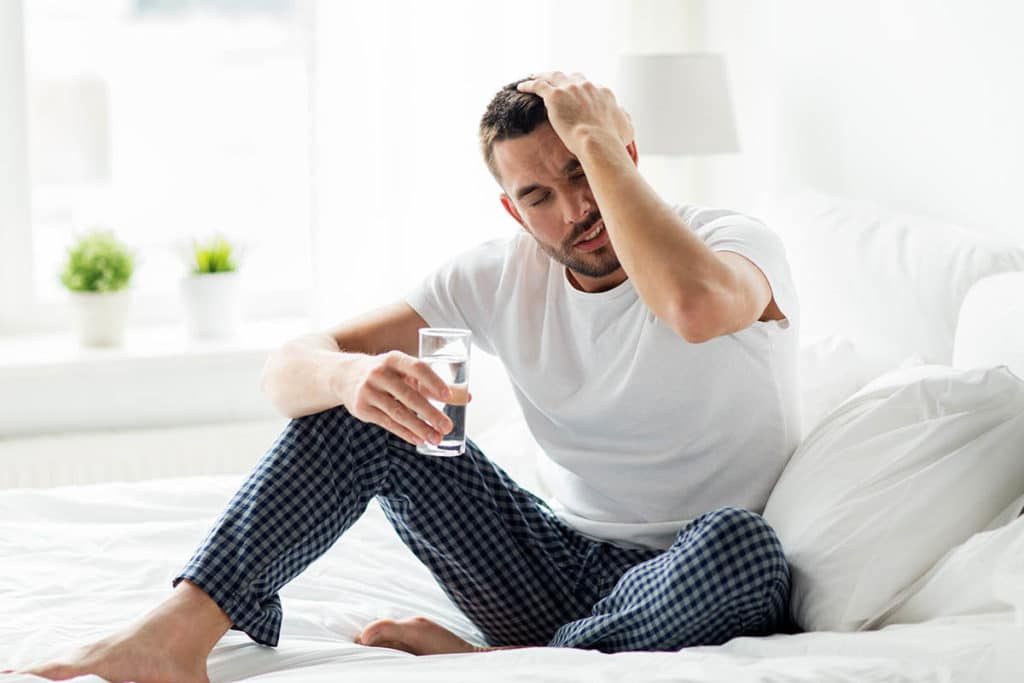 a person sits holding their head with a glass of water possibly struggling with Signs of alcohol withdrawals