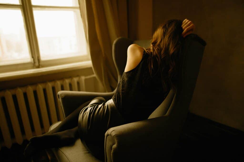 A woman is resting her head on the back of a chair, with one arm covering her face. She is seated in a cozy, dimly lit room near a window, with natural light filtering through.